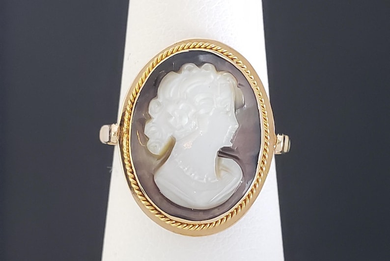 May include: Gold ring featuring an oval cameo with a white profile against a dark background. The cameo is framed by a twisted gold rope design. The ring is set on a white stand against a dark background.