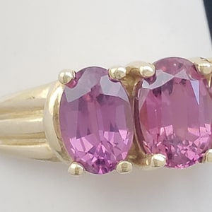 May include: A gold ring featuring three oval-cut pink gemstones. The gemstones are set in a row, with a gold band. The ring is displayed on a white surface against a dark blue background.