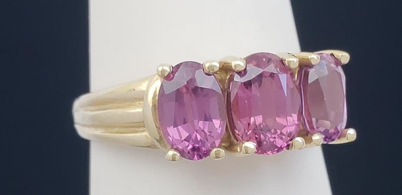 May include: A gold ring featuring three oval-cut pink gemstones. The gemstones are set in a row, with a gold band. The ring is displayed on a white surface against a dark blue background.