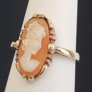 May include: A gold ring with a cameo featuring a woman's profile in a white cameo set in a peach-colored shell.