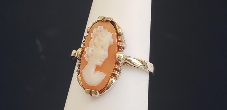 May include: A gold ring with a cameo featuring a woman's profile in a white cameo set in a peach-colored shell.