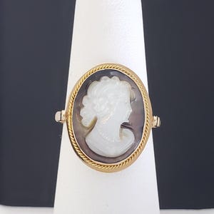 May include: A gold ring featuring an oval cameo with a white profile against a dark background. The ring has a twisted rope-like border and is displayed on a white stand, showcasing the jewelry's design and craftsmanship.