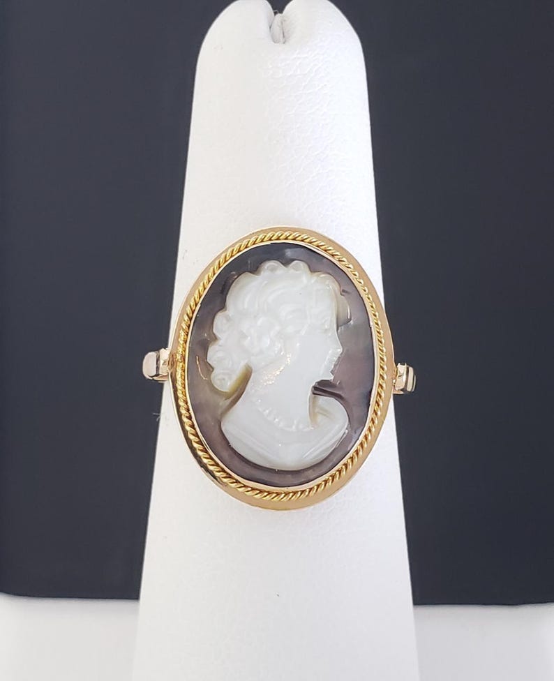 May include: A gold ring featuring an oval cameo with a white profile against a dark background. The ring has a twisted rope-like border and is displayed on a white stand, showcasing the jewelry's design and craftsmanship.