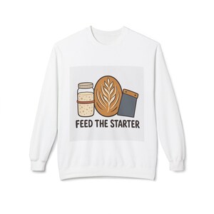 May include: A white long-sleeve sweatshirt featuring a graphic of a glass jar of sourdough starter, a loaf of bread with a wheat stalk score, and a grey dough scraper. Below the illustration, the text reads FEED THE STARTER.