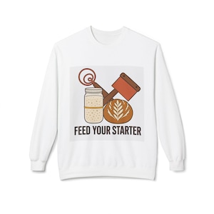 May include: A white long-sleeve sweatshirt featuring a graphic of a glass jar with sourdough starter, a whisk, a bread lame, and a round loaf of bread. Below the illustration, the text reads FEED YOUR STARTER in bold black capital letters.