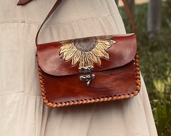 May include: A brown leather crossbody bag with a sunflower design and a metal clasp. The bag has a brown leather strap and is worn over a white dress.
