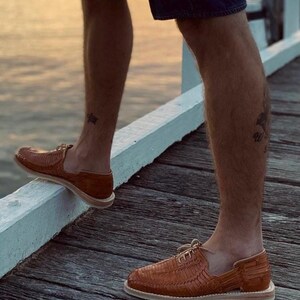 May include: A pair of brown leather sandals with woven straps and a closed toe. The sandals have a white sole and are worn by a person standing on a wooden deck.