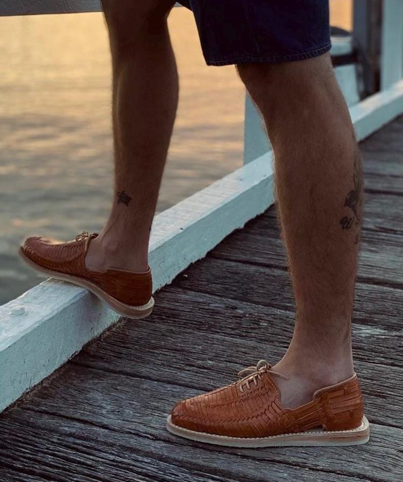 May include: A pair of brown leather sandals with woven straps and a closed toe. The sandals have a white sole and are worn by a person standing on a wooden deck.