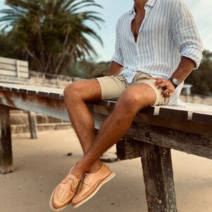 May include: A person wearing a light blue striped shirt, khaki shorts, and brown leather sandals with woven details. The person is sitting on a wooden platform with their legs dangling over the edge. The background is a beach with sand and palm trees.