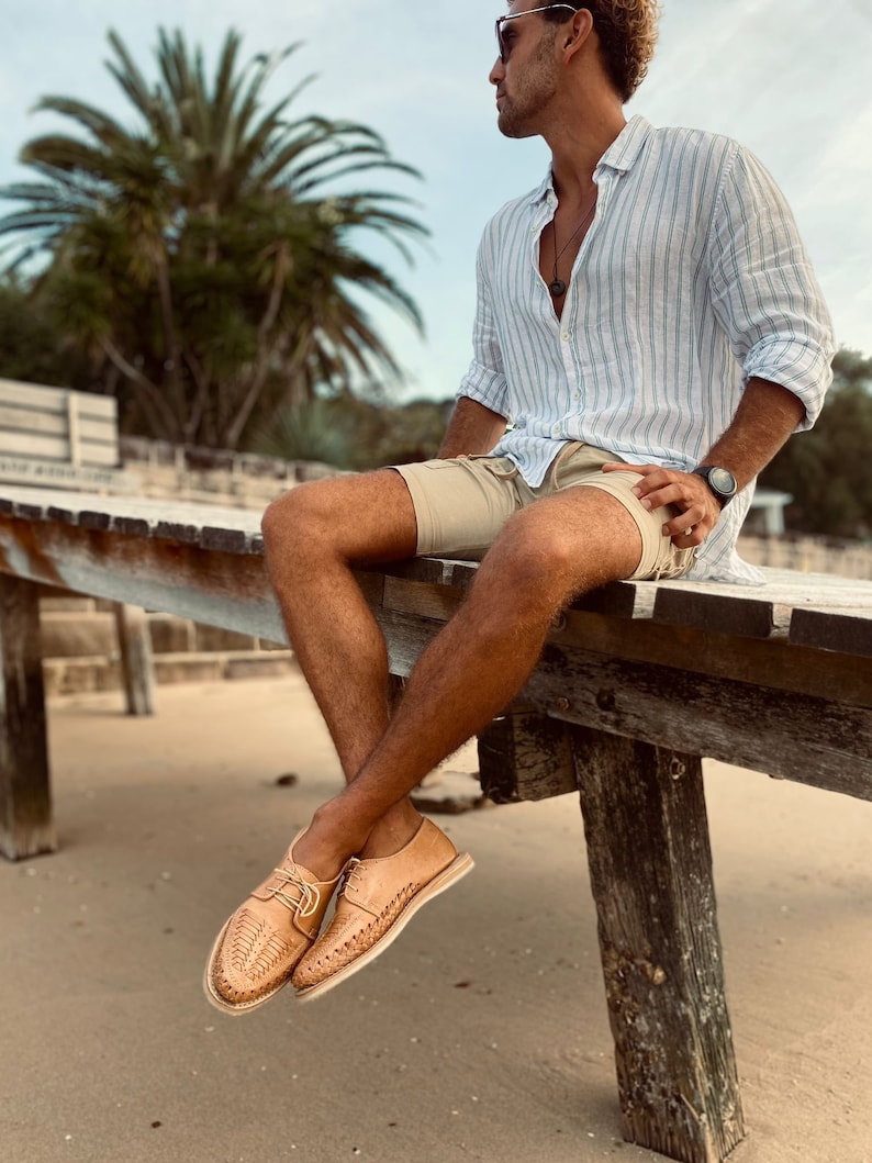 May include: A person wearing a light blue striped shirt, khaki shorts, and brown leather sandals with woven details. The person is sitting on a wooden platform with their legs dangling over the edge. The background is a beach with sand and palm trees.