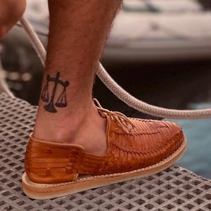 May include: A pair of tan leather sandals with a woven design. The sandals have a lace-up closure and are worn on a person's foot. A tattoo of a scale is visible on the person's ankle.