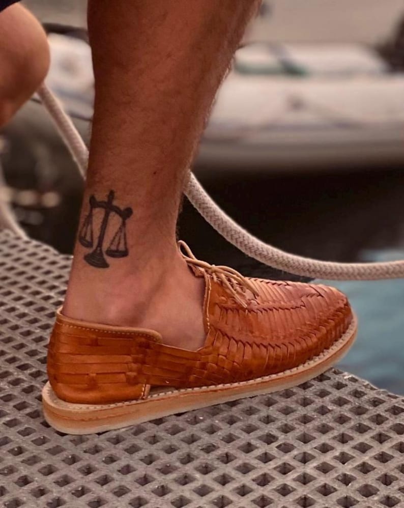 May include: A pair of tan leather sandals with a woven design. The sandals have a lace-up closure and are worn on a person's foot. A tattoo of a scale is visible on the person's ankle.