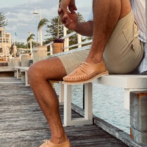 May include: A person wearing tan leather sandals with woven detailing. The sandals have a lace-up closure and are worn on a wooden dock.
