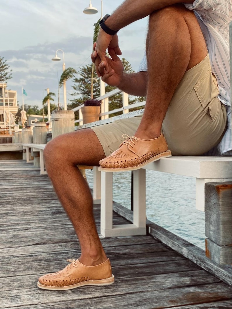 May include: A person wearing tan leather sandals with woven detailing. The sandals have a lace-up closure and are worn on a wooden dock.