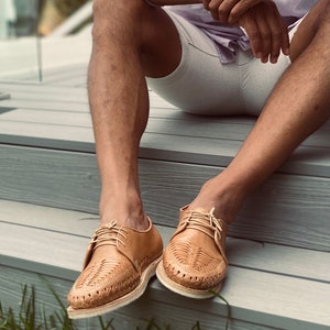 May include: A pair of tan leather shoes with woven detailing. The shoes have laces and are worn by a person sitting on a wooden deck.