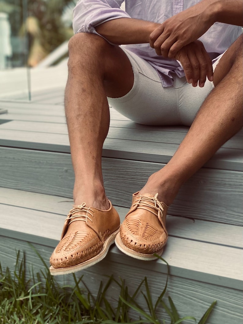 May include: A pair of tan leather shoes with woven detailing. The shoes have laces and are worn by a person sitting on a wooden deck.