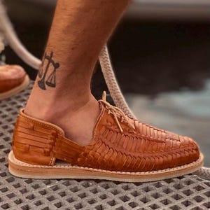 May include: Tan leather sandals with a woven design and a lace-up closure. The sandals have a thick sole and are worn on a person's feet.
