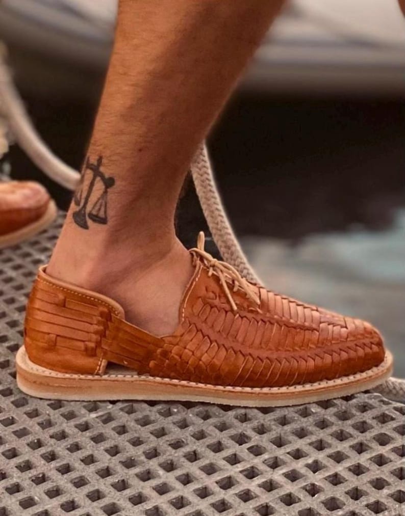 May include: Tan leather sandals with a woven design and a lace-up closure. The sandals have a thick sole and are worn on a person's feet.