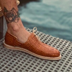 May include: A pair of brown leather woven sandals with a lace-up closure. The sandals are on a light gray textured surface.