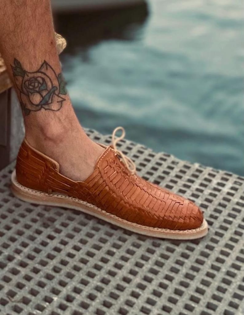 May include: A pair of brown leather woven sandals with a lace-up closure. The sandals are on a light gray textured surface.