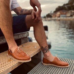 May include: A pair of brown leather sandals with a woven design. The sandals are worn by a person sitting on a wooden dock.