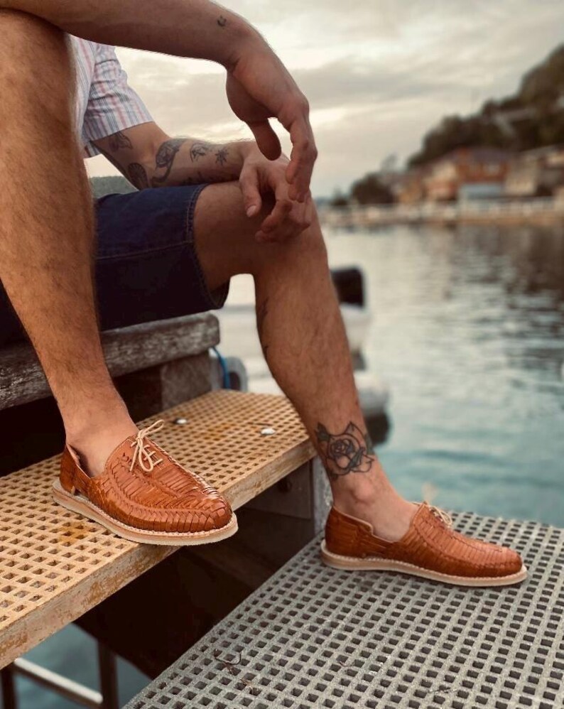 May include: A pair of brown leather sandals with a woven design. The sandals are worn by a person sitting on a wooden dock.
