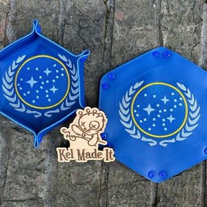 May include: Two blue hexagonal dice trays with a white and yellow embroidered Starfleet logo. The trays are on a brick surface with a wooden sign that says "Kel Made It".