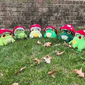 May include: Seven plush frog toys with red mushroom caps and white polka dots are arranged on green grass. The frogs have various shades of green bodies, yellow bellies, and expressive eyes. Fallen leaves are scattered around the toys.