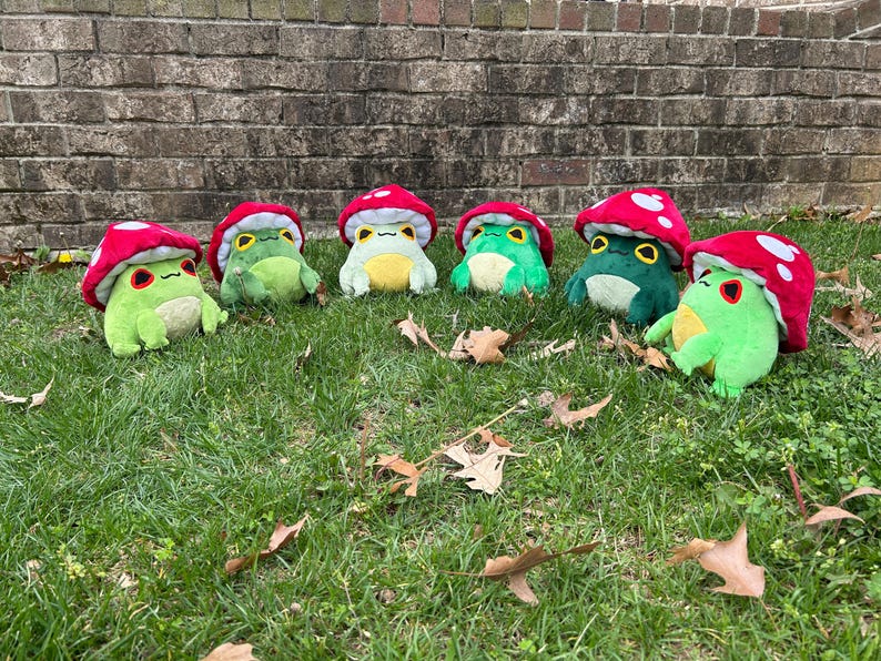 May include: Seven plush frog toys with red mushroom caps and white polka dots are arranged on green grass. The frogs have various shades of green bodies, yellow bellies, and expressive eyes. Fallen leaves are scattered around the toys.