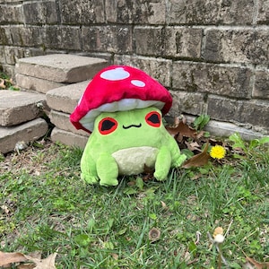 May include: A plush frog wearing a red mushroom hat with white spots. The frog is green with a cream-colored belly and black eyes with red accents. It is sitting on green grass with a brick wall in the background.