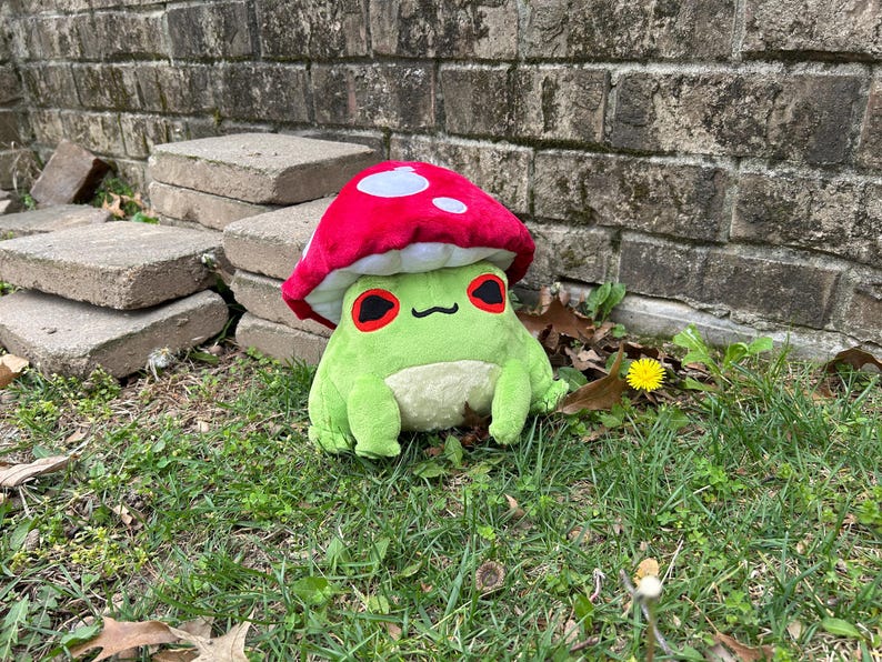 May include: A plush frog wearing a red mushroom hat with white spots. The frog is green with a cream-colored belly and black eyes with red accents. It is sitting on green grass with a brick wall in the background.