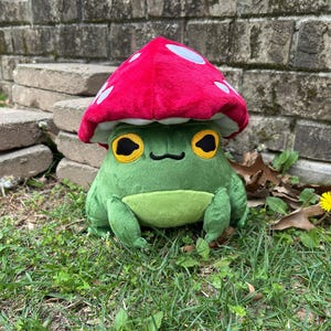 May include: A plush green frog wearing a red mushroom cap with white spots. The frog has large yellow eyes with black pupils and a small black smile. The frog is sitting on green grass.