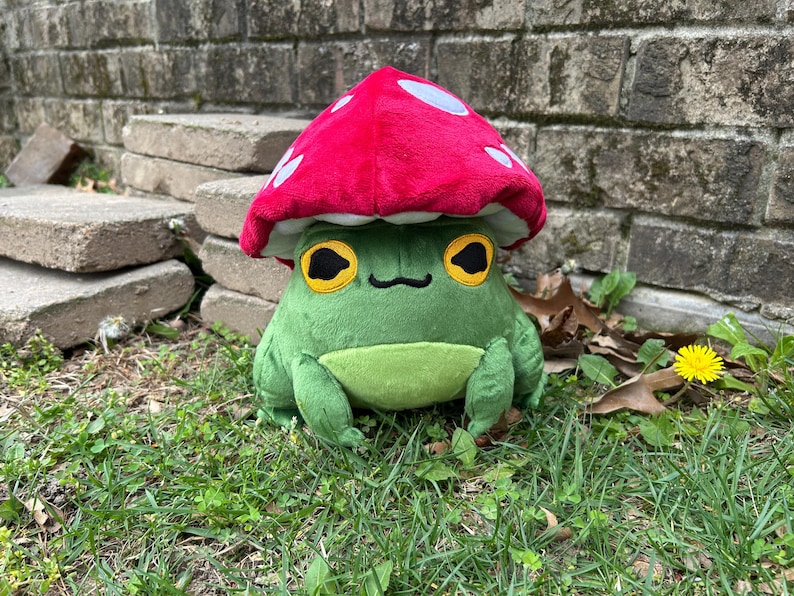 May include: A plush green frog wearing a red mushroom cap with white spots. The frog has large yellow eyes with black pupils and a small black smile. The frog is sitting on green grass.