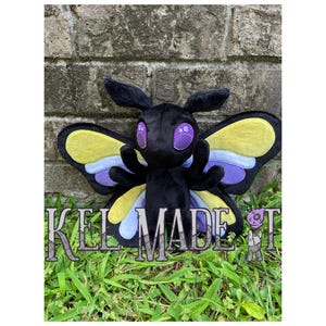 May include: A plush moth toy with a black body, purple eyes, and large wings in yellow, blue, and purple. The text "KEL MADE IT" is across the bottom. The toy is set against a brick wall and grass.