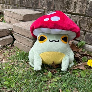 May include: A plush frog wearing a red mushroom hat with white spots. The frog is light green with a yellow belly and large, yellow-rimmed eyes. It is sitting on grass near a brick wall.