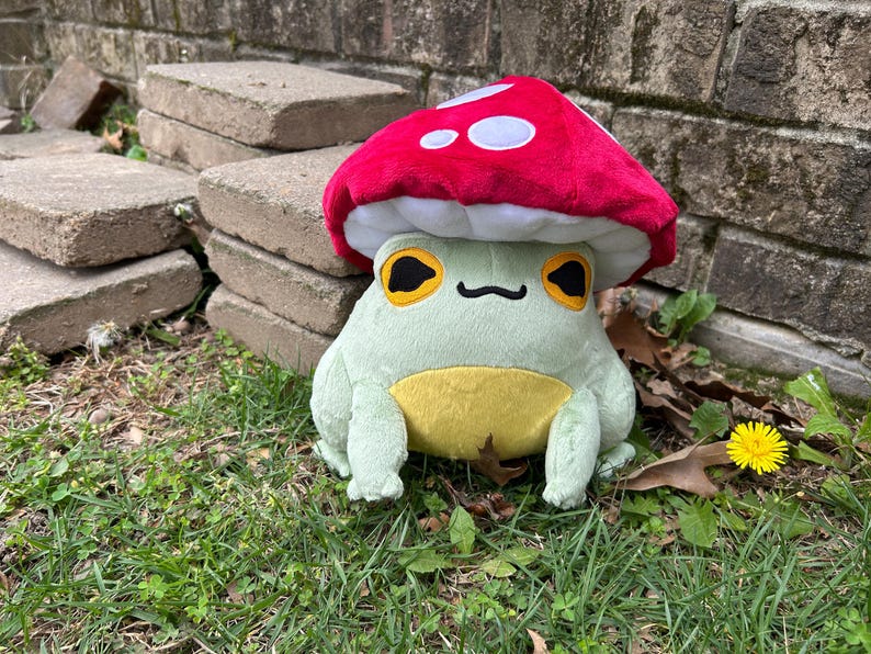 May include: A plush frog wearing a red mushroom hat with white spots. The frog is light green with a yellow belly and large, yellow-rimmed eyes. It is sitting on grass near a brick wall.