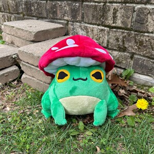 May include: A plush green frog wearing a red mushroom hat with white spots. The frog has large yellow eyes with black pupils and a small black mouth. The frog is sitting on green grass.
