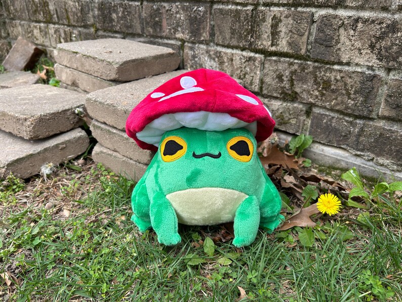 May include: A plush green frog wearing a red mushroom hat with white spots. The frog has large yellow eyes with black pupils and a small black mouth. The frog is sitting on green grass.
