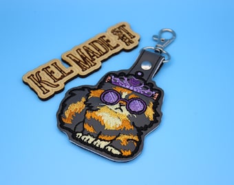 May include: A keychain featuring an embroidered patch of a fluffy orange and gray cat wearing purple round glasses and a purple crown, attached to a silver metal clasp. Beside it is a wooden tag with the text KEL MADE engraved in black.