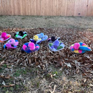May include: A collection of colorful plush snake toys in various colors, including pink, blue, yellow, and green. The stuffed animals are coiled on a bed of fallen leaves, with a wooden fence in the background.