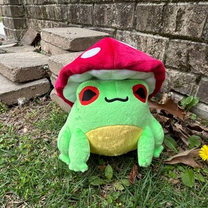 May include: A plush green frog with a yellow belly and red eyes, wearing a red mushroom cap with white spots. The frog is sitting on grass near a brick wall and concrete blocks.
