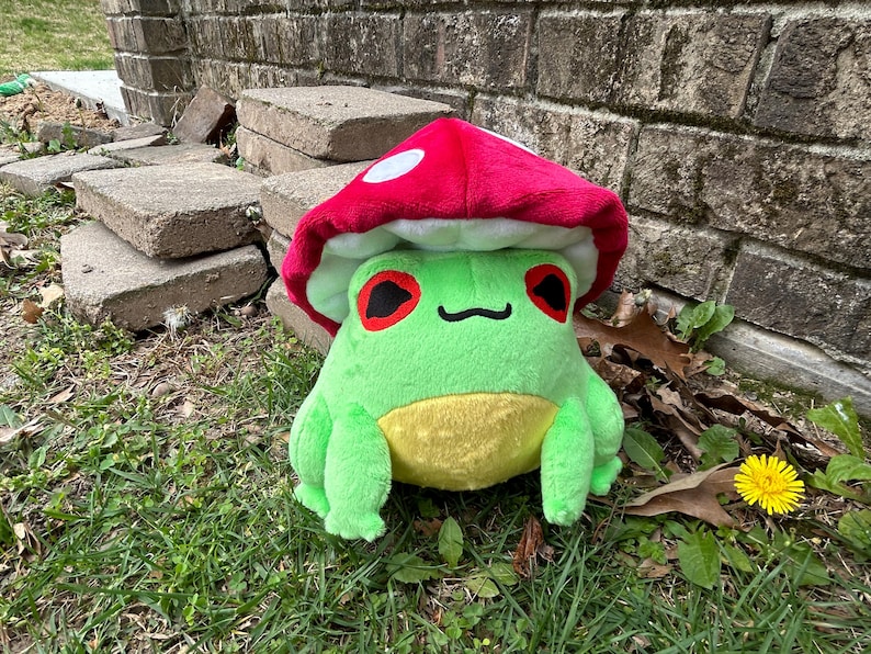 May include: A plush green frog with a yellow belly and red eyes, wearing a red mushroom cap with white spots. The frog is sitting on grass near a brick wall and concrete blocks.