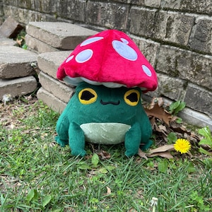 May include: A plush frog wearing a red mushroom hat with white spots. The frog is green with yellow eyes and a cream-colored belly. The plush toy is sitting on grass near a brick wall and concrete blocks.