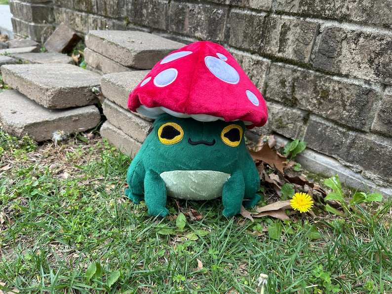 May include: A plush frog wearing a red mushroom hat with white spots. The frog is green with yellow eyes and a cream-colored belly. The plush toy is sitting on grass near a brick wall and concrete blocks.