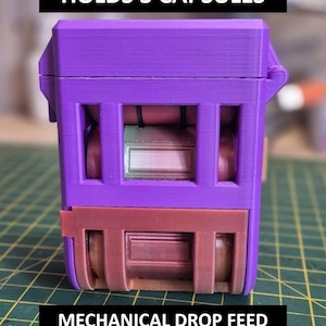 May include: A purple and brown mechanical drop feed device. The device has a rectangular shape with three visible compartments. The text "HOLDS 3 CAPSULES" is at the top, and "MECHANICAL DROP FEED" is at the bottom.