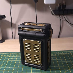 May include: A black rectangular box with brass-colored accents. The box features a hinged lid and a front panel with horizontal vents. The design incorporates brass-colored plates and screws, giving it an industrial aesthetic.