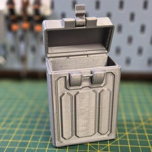 May include: A small, gray, rectangular box with a hinged lid. The box has a textured surface and a latch on the front. The lid is open, revealing the empty interior of the box.