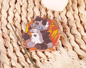 Op de afbeelding: Een grijze en witte kat met blauwe ogen die een rode en oranje vlammen-emaille pin draagt. De kat kijkt naar links en heeft een serieuze uitdrukking.
