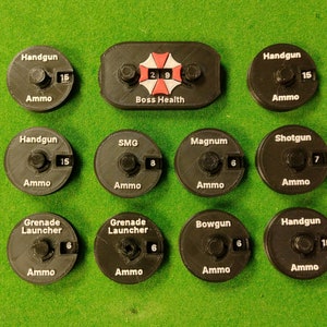 May include: A set of 10 black plastic tokens with white text and numbers. The tokens are for a board game and show the amount of ammo for different weapons, including Handgun, SMG, Magnum, Shotgun, Grenade Launcher, and Bowgun. One token shows a red and white umbrella symbol with the text "Boss Health" and the numbers "29".