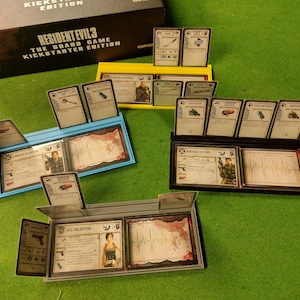 Può includere: Tre porta carte di diversi colori per il gioco da tavolo Resident Evil 3. I porta carte sono in plastica e hanno slot per le carte. I porta carte sono etichettati con i nomi dei personaggi: Jill Valentine, Carlos Oliveira e Mikhail Victor. I porta carte sono su una superficie di feltro verde.